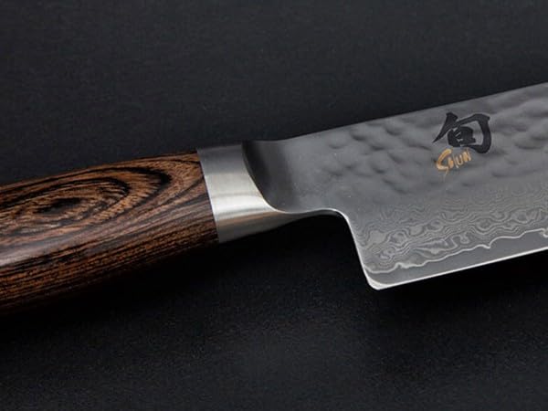 KAI Shun Premier Tim Mälzer Santoku mit 18 cm Klinge. VG-MAX Stahlkern (61 HRC), 32 Lagen Damast, hochglanzpoliert mit Hammerschlagoberfläche. Griff aus gemasertem Pakkaholz in Mittelbraun. Das Präzisionswerkzeug aus Japan.